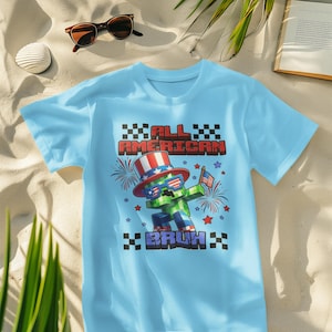 May include: Light blue t-shirt with a graphic featuring a pixelated character wearing a patriotic hat and sunglasses, holding a flag. The text reads "ALL AMERICAN BRUH" in red and black.