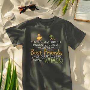Puede incluir: Camiseta gris oscuro con el texto "TURTLES ARE GREEN DUCKS GO QUACK WE'RE Best Friends CAUSE OUR HEADS ARE EQUALLY WHACK!" con un pato y una tortuga de dibujos animados. Gafas de sol, una bebida y un libro en el fondo.