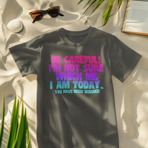 Op de afbeelding: Donkergrijs t-shirt met de tekst "BE CAREFUL! I'M NOT SURE WHICH ME I AM TODAY. YOU HAVE BEEN WARNED" in roze, paars en blauw. Het t-shirt ligt op zand, met een zonnebril en een drankje.