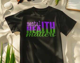 May include: Black t-shirt with the text "mental HEALTH matters" in purple and green. The shirt is displayed on a sandy surface with sunglasses, a drink, a seashell, and a book. The shirt is a casual wear item.