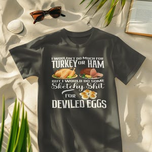 Puede incluir: Camiseta gris oscuro con el texto "I wouldn't do much for turkey or ham but I would do some sketchy shit for deviled eggs." La camiseta presenta imágenes de pavo, jamón y huevos rellenos. Gafas de sol, una bebida y un libro están en el fondo.