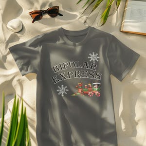 May include: A gray t-shirt with the words "BIPOLAR EXPRESS" in a stylized font, accompanied by snowflake and train graphics. The train is red, green, and white. The shirt is displayed on a sandy surface with sunglasses and a drink.