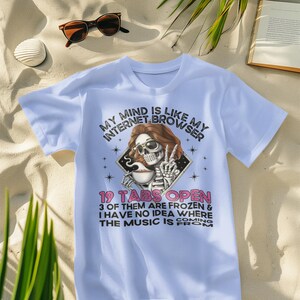 以下が含まれることがあります： 薄い青色のTシャツに、サングラスをかけてコーヒーカップを持った骸骨が描かれています。Tシャツには「My mind is like my internet browser 19 tabs open 3 of them are frozen & I have no idea where the music is coming from.」という文字が書かれています。