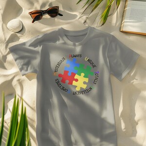 Puede incluir: Una camiseta gris con un diseño de pieza de rompecabezas de colores y las palabras "Always Unique Totally Intelligent Sometimes Mysterious" alrededor de la pieza del rompecabezas.