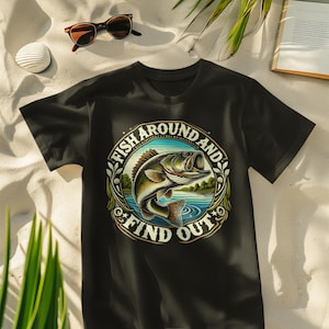 Pode incluir: Camiseta preta com um gráfico circular apresentando um peixe saltando da água, rodeado por folhagens. O texto "FISH AROUND AND FIND OUT" está arqueado ao redor do peixe. Óculos de sol, bebida e livro ao fundo.