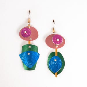 May include: A pair of handmade earrings with a unique, abstract design. Each earring features a pink oval shape with a purple circle, a green and blue geometric shape, and a gold-colored stem. The earrings are suspended from silver hooks.