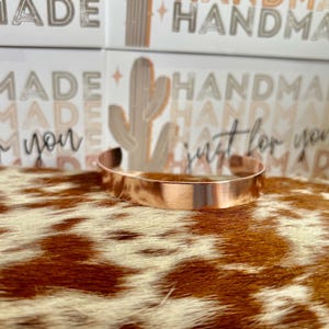 May include: A copper cuff bracelet with a brushed metal finish. The bracelet is displayed on a cowhide-patterned surface, with cards in the background that read "MADE HANDMADE" and "just for you".