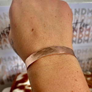 May include: A hammered copper cuff bracelet. The bracelet has a textured surface and a warm, reddish-brown colour. The cuff is a simple, minimalist design, suitable for everyday wear. The bracelet is approximately 2 cm wide.