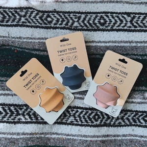 May include: Three Wild One Twist Toss dog toys in peach, navy, and pink, each on a tan card with the text "Wild One Twist Toss Throw Toy For Dogs". The toys are star-shaped and made of rubber.
