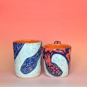 on white speckle clay, I have carved an image of a two headed snake design of mine- the inside color is bright orange, just like the eyes of the snakes, and the snakes are dark blue. they're scales/flame markings are colored with a mixture of different oranges handle is pinched with blue glaze and orange scale textures in the pinched parts----- image of my cup and mug next to each other