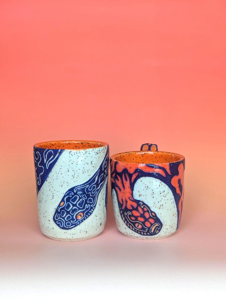 on white speckle clay, I have carved an image of a two headed snake design of mine- the inside color is bright orange, just like the eyes of the snakes, and the snakes are dark blue. they're scales/flame markings are colored with a mixture of different oranges handle is pinched with blue glaze and orange scale textures in the pinched parts----- image of my cup and mug next to each other
