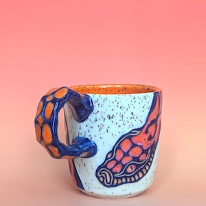 on white speckle clay, I have carved an image of a two headed snake design of mine- the inside color is bright orange, just like the eyes of the snakes, and the snakes are dark blue. they're scales/flame markings are colored with a mixture of different oranges handle is pinched with blue glaze and orange scale textures in the pinched parts