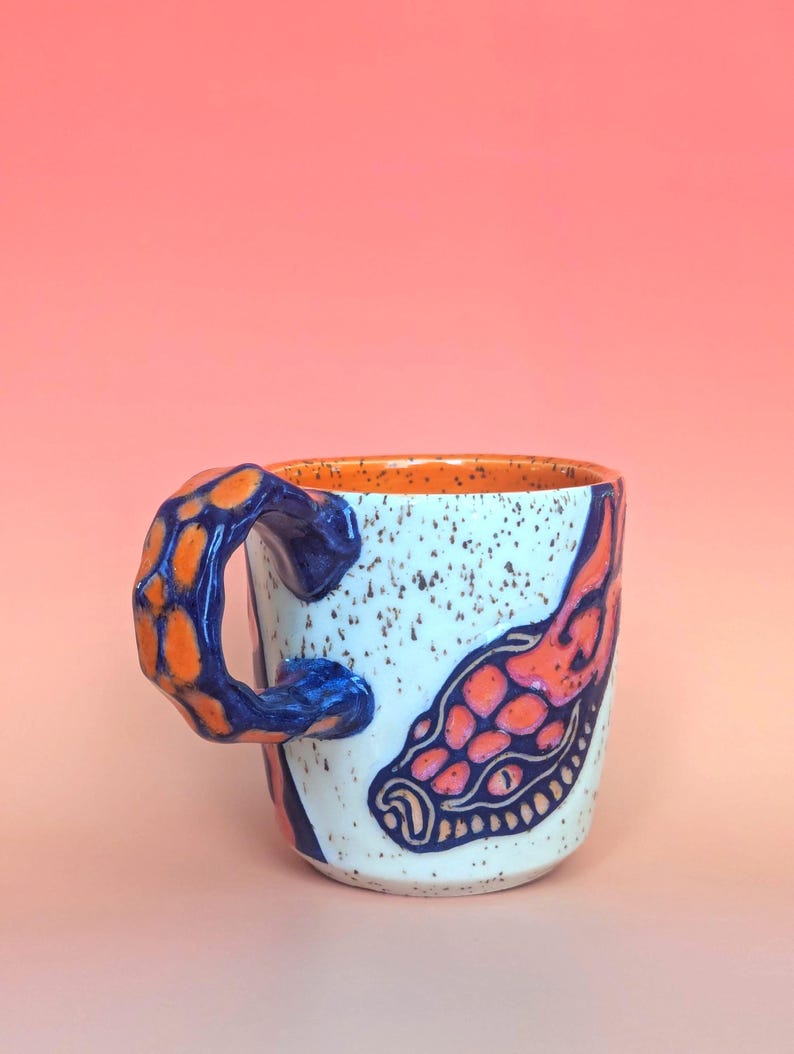 on white speckle clay, I have carved an image of a two headed snake design of mine- the inside color is bright orange, just like the eyes of the snakes, and the snakes are dark blue. they're scales/flame markings are colored with a mixture of different oranges handle is pinched with blue glaze and orange scale textures in the pinched parts