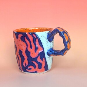 on white speckle clay, I have carved an image of a two headed snake design of mine- the inside color is bright orange, just like the eyes of the snakes, and the snakes are dark blue. they're scales/flame markings are colored with a mixture of different oranges handle is pinched with blue glaze and orange scale textures in the pinched parts