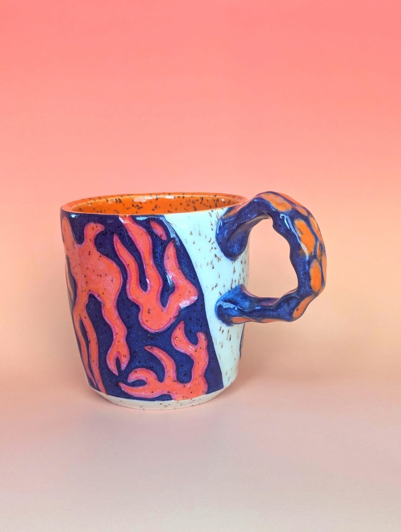 on white speckle clay, I have carved an image of a two headed snake design of mine- the inside color is bright orange, just like the eyes of the snakes, and the snakes are dark blue. they're scales/flame markings are colored with a mixture of different oranges handle is pinched with blue glaze and orange scale textures in the pinched parts