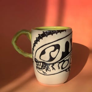 May include: Cream-colored ceramic mug with a black dinosaur skull design. The mug has a light green handle and a yellow interior rim. The design is a stylized illustration of a dinosaur skull. The mug is a unique and artistic piece.