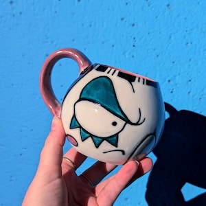 May include: A white ceramic mug with a cartoon face design. The mug features a teal and black illustration of a face with a pink handle. The mug is held in a hand against a blue background.