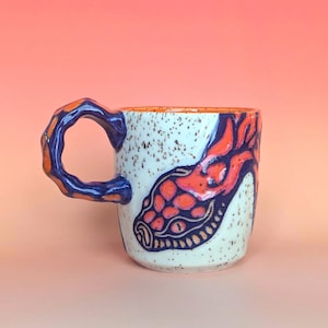 May include: A ceramic mug with a speckled off-white glaze featuring a hand-painted orange and dark blue snake design. The mug has a thick, textured, irregular handle in matching blue and orange colors and a bright orange interior finish.