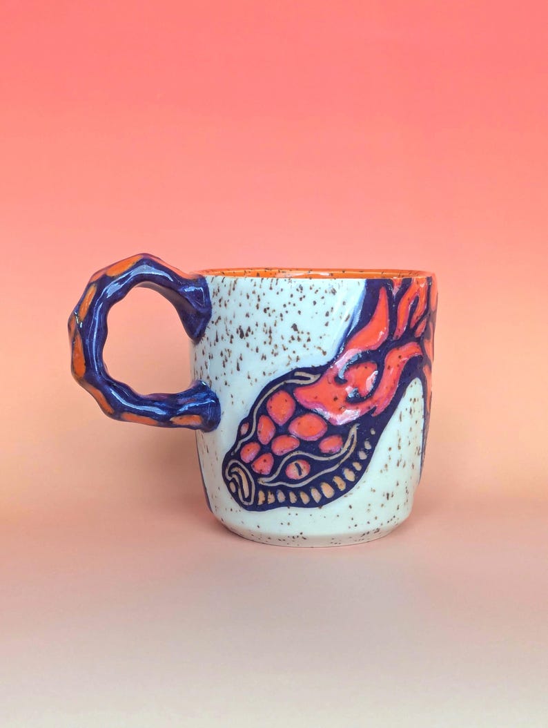 on white speckle clay, I have carved an image of a two headed snake design of mine- the inside color is bright orange, just like the eyes of the snakes, and the snakes are dark blue. they're scales/flame markings are colored with a mixture of different oranges