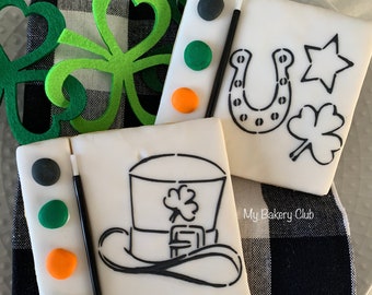 Puede incluir: Dos galletas de azúcar decoradas con diseños para pintar. Una galleta tiene un sombrero de duende, la otra tiene una herradura, un trébol y una estrella. Ambas galletas tienen una paleta de pintura con cuatro puntos de pintura y un pincel.