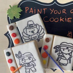 May include: Three paint-your-own cookies with Thanksgiving themes. The cookies are decorated with a turkey, a Native American girl, and a paint palette with paintbrushes. The cookies are on a wooden surface with a chalkboard sign that says "Paint Your Own Cookie".