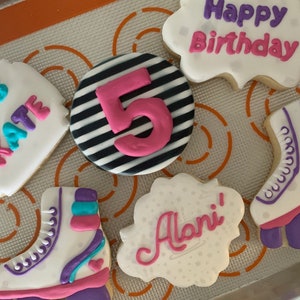 May include: Five decorated sugar cookies with a roller skate theme. One cookie says "Let's Skate" in pink and purple lettering. Another cookie has black and white stripes with a pink number 5 in the center. A third cookie says "Happy Birthday" in purple and pink lettering. A fourth cookie says "Alani!" in pink lettering. The fifth cookie is a white roller skate with purple and blue accents.