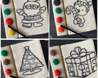May include: Four Christmas-themed paint-your-own cookies with edible paint. Each cookie features a different design: Santa Claus, a snowman, a Christmas tree, and a gift box. The cookies are decorated with edible paint pots in various colours.