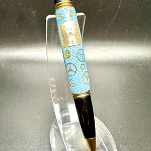 May include: A blue and gold pen with a clock face design and gears. The pen is on a clear plastic stand.