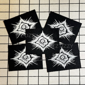 May include: A collection of five black square patches featuring a white, stylized eye design with radiating lines. The patches have a gothic or occult theme, with the text "TZDZZY" visible on some.