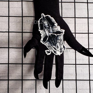 May include: A black and white sticker featuring a haunted house design. The sticker is held by a black gloved hand with long, pointed fingernails. The background is a grid pattern.