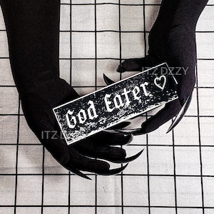May include: A black and white rectangular sign with the text "God Eater" in a gothic font, held by black-gloved hands with long, sharp claws. A white heart is next to the text. The background is a checkered pattern.