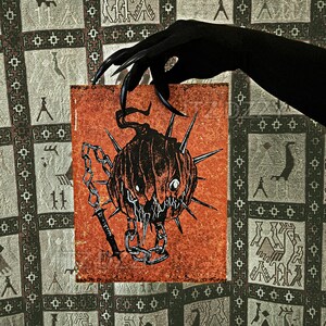 May include: A square print featuring a menacing pumpkin with sharp spikes, a chain, and a blade. The pumpkin is black and orange, with a sinister expression. The print is held by a black-gloved hand against a patterned background.