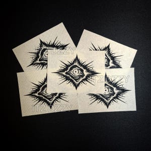 May include: A collection of off-white fabric patches featuring a black eye design within a starburst shape. The patches are arranged on a dark surface, with the text "ITZ UZZY" visible on some.