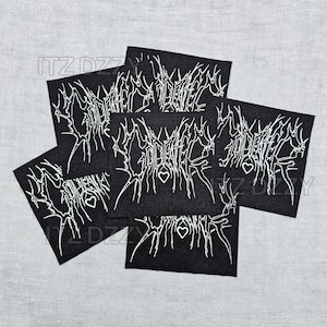 May include: A collection of black fabric patches with white, stylized text and heart symbols. The patches are arranged in a slightly overlapping pattern, with the text appearing to be a band or brand name. The background is a light gray fabric.