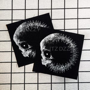 May include: Two black square patches with a white skull design. The skulls have detailed features and are surrounded by a white, circular pattern. The text "ITZ DZZY" is visible on one patch.