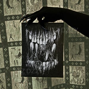 May include: A black and white illustration of a skull with open jaws, held by a gloved hand with long, sharp claws. The artwork is on a rectangular black canvas. The background features a patterned fabric.