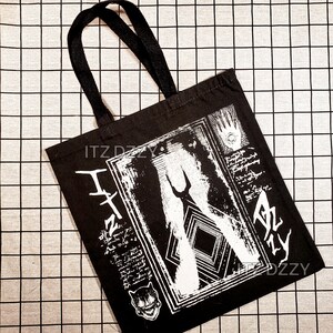 May include: Black tote bag with black handles. The bag features a white graphic print with abstract shapes, text, and a cat illustration. The text "ITZ DZZY" is printed on the bag.