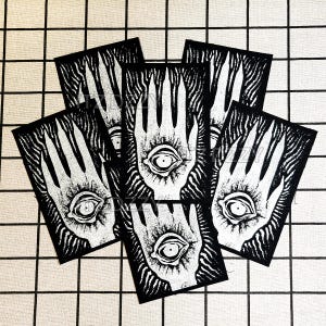 Op de afbeelding: Een verzameling van zes zwart-witte patches. Elke patch toont een gestileerde hand met een oog in de palm. De patches zijn gerangschikt op een geruit patroon.