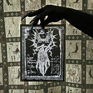 May include: A black and white art print with a gothic design. The print features a central illustration of a creature with a single eye, surrounded by thorns and script. The print is held by a black-gloved hand with long, pointed nails.