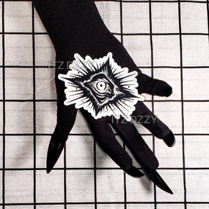 May include: A black and white sticker featuring an eye design within a starburst shape. The sticker is placed on a black surface. The sticker has a detailed illustration of an eye with radiating lines.