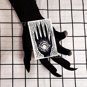 May include: A black and white rectangular card featuring a stylized eye within a hand design. The card is held against a black background. The design has a mystical or occult theme.