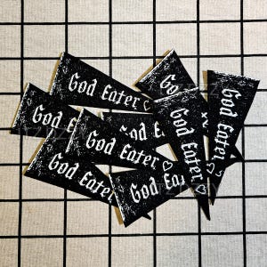 May include: A collection of black triangular pennants with the words "God Eater" printed in white. The pennants are arranged in a scattered pattern against a checkered background. Some pennants have small heart symbols.