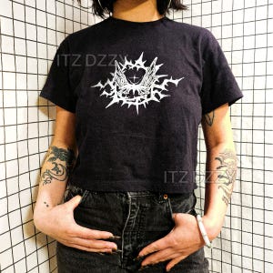 May include: Black crop top with a white graphic design featuring a stylized winged creature with spiky details. The text "ITZ DZZZ" is printed on the shirt. The model is wearing black jeans.
