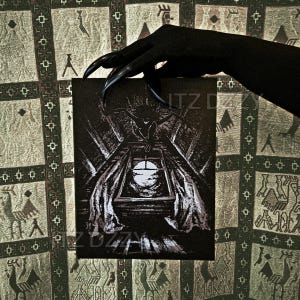 May include: A black and white illustration of a creature perched above a window, with the text "ITZ DZZY" and "ZDZZY" on the card. The image is held by a black hand with long, pointed nails. The background is a patterned fabric.