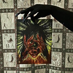 May include: A dark, stylized illustration of a screaming face with sharp teeth and hands covering the face. The artwork is held by a black hand with long, pointed nails. The background is a patterned fabric.
