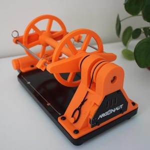 May include: An orange 3D-printed mechanical device with two large, spoked wheels and a black base. The device has a complex structure with various gears and supports. The text "ARGONAUT" is visible on the side.