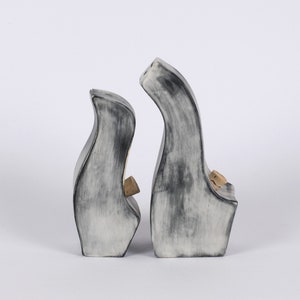 Puede incluir: Dos esculturas de cerámica en un esmalte gris y blanco. Las esculturas son abstractas y tienen una forma redondeada y orgánica. Ambas están de pie y están colocadas una al lado de la otra.