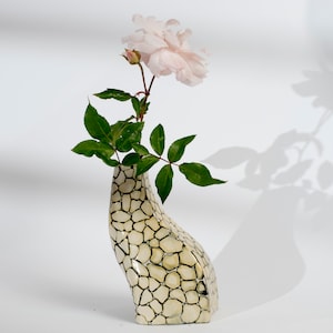 Könnte beinhalten: Eine weiße Keramikvase mit strukturierter Oberfläche, in Form einer Hand, die eine einzelne rosa Rose mit grünen Blättern hält.