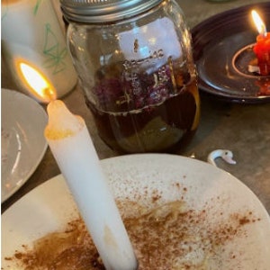 May include: A lit white candle on a white plate with brown powder. A glass jar with a metal lid contains a dark liquid and pink petals. Other candles and jars are in the background.