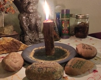 May include: A lit candle on a blue plate with a stone base, surrounded by stones with etched text. A gargoyle statue with a purple crown sits behind the candle. Other items include jars and bottles.